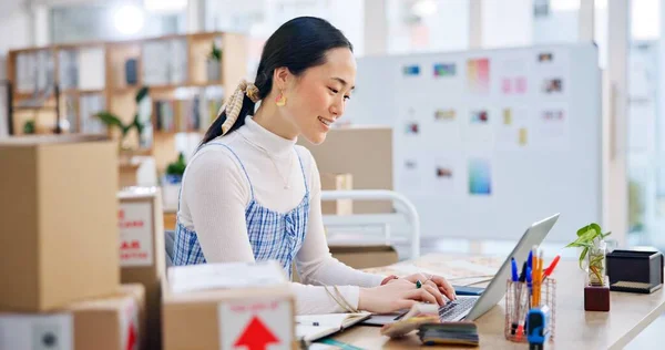 Ecommerce, Asian woman at laptop with typing and smile for sales report and work at fashion startup. Online shopping, boxes and small business owner with happiness, computer and website shop at desk