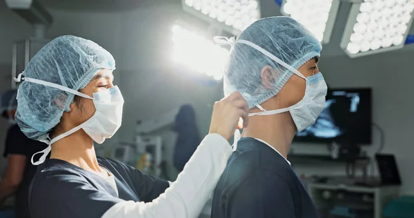 Surgery, hospital and doctors tie mask in operating room for medical service, preparation and operation. Healthcare team, safety scrubs and people with uniform for emergency, procedure and protection.