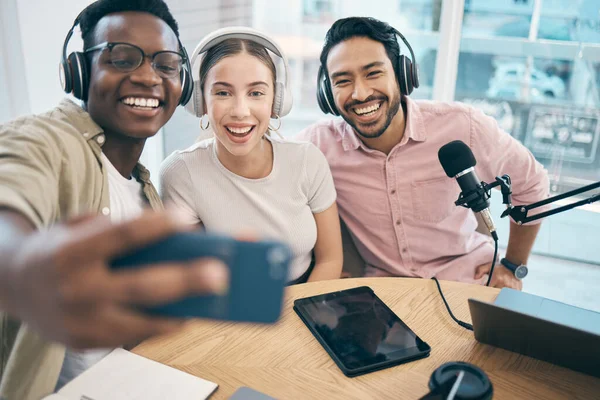 Podcast, smile and group selfie of friends together bonding, live streaming and people recording broadcast in studio. Happy team of radio hosts take picture at table for social media or collaboration.