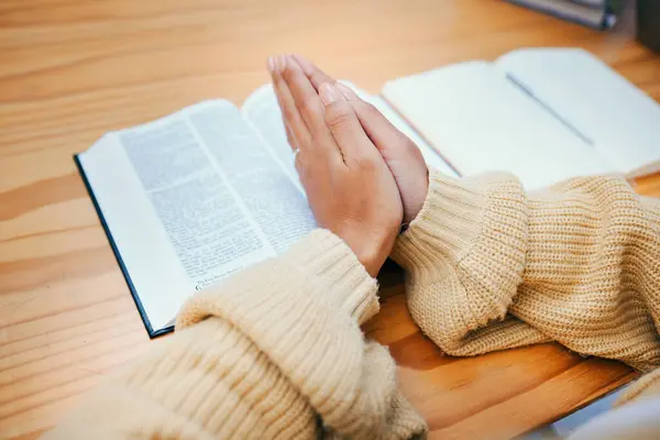 Hands, bible and praying at desk, religion and Christian worship in home at table. Closeup, holy book and woman in meditation for God, Jesus and Christ for faith in spiritual gospel, praise or hope.