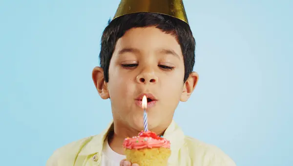 Blowing candle, child and a cupcake for a birthday, celebration or a wish on a studio background. Happy, face portrait and a hungry boy kid with party food, cake or dessert for a present or event.