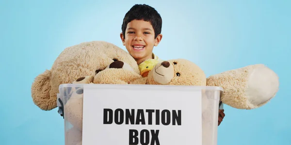 Boy in studio with toys, donation in box and smile for children, social charity and hope in kindness. Care, donate and happy kindergarten child with teddy bear package at kids ngo on blue background