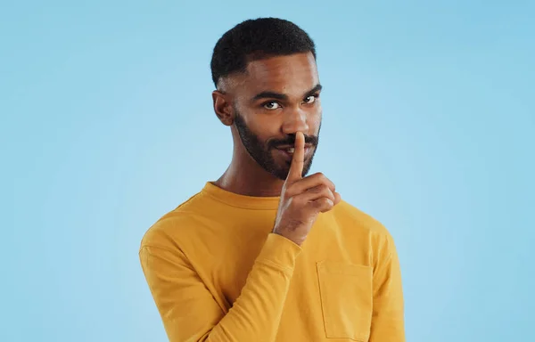 Secret, face and man with finger on lips in studio for quiet, privacy or hush news on blue background. Whisper, drama and portrait of guy model with confidential hand emoji for gossip or announcement.