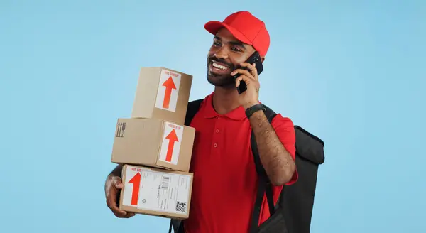 Man, logistics and courier with cellphone in communication to client, delivery or boxes in studio. Indian person, supply chain and cargo in conversation with customer, packages and blue background.