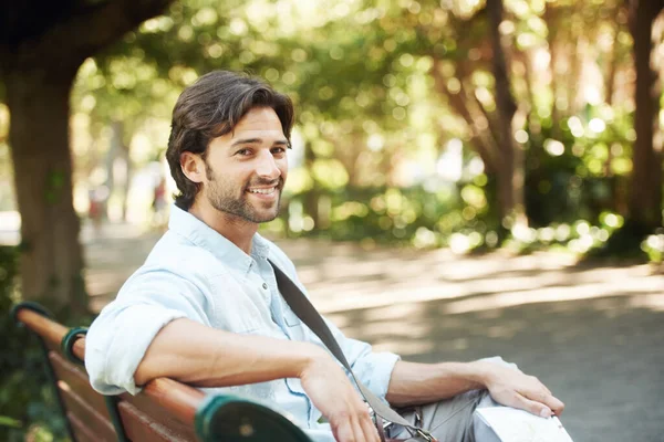 Relax, portrait or happy man in park on a bench in summer for fresh air, holiday vacation or break in garden. Confident, smile or calm person in nature resting or waiting in a natural environment.