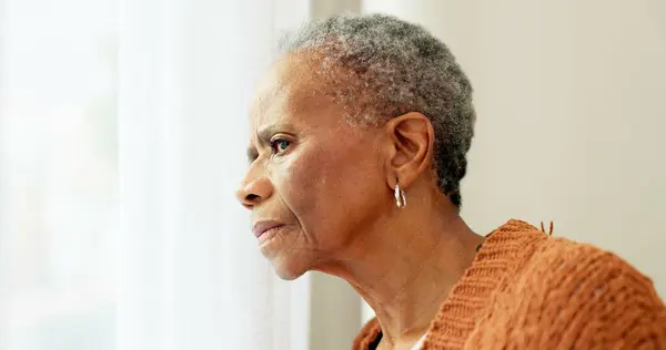 Senior woman, face and thinking by window at home to remember memory and relax in retirement. Serious black elderly person or old lady at nursing facility with hope, sad emotion and Alzheimer disease.