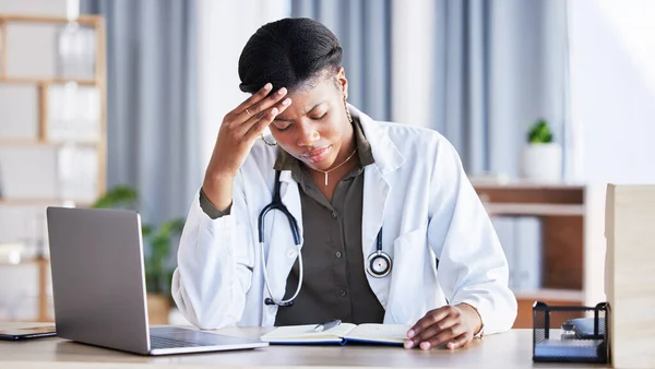 Doctor, African woman and headache with laptop at desk, clinic office or stress with thinking at job. Overworked medic, fatigue and burnout in hospital, book or anxiety for mental health by computer.