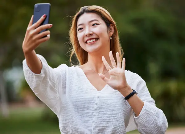 Happy, woman and wave hello to phone for video call, blog or social media, communication or post on internet in the park. Selfie, face and girl with a smile for smartphone conversation or film.