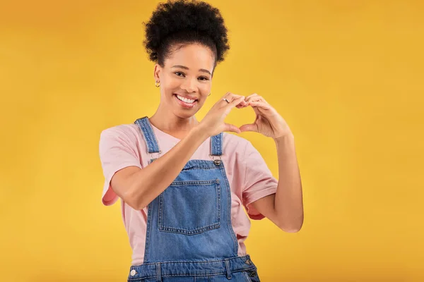 Black woman, hands and sign for a heart, love and portrait of care or support on yellow background in studio. Happy, face and girl with hand gesture for loving, kindness or emoji for caring mindset.