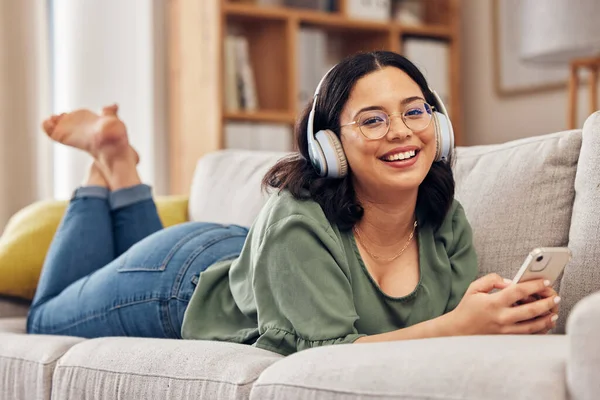 Woman, portrait and headphones with smartphone on sofa, for hearing radio, online subscription and podcast. Happy female person listening to music, streaming audio and sound on mobile in living room.