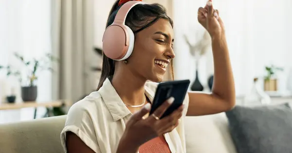 Phone, music and happy woman dance on sofa in home, singing and listening to audio. Smartphone, excited Indian person on headphones and radio, sound or freedom in living room on mobile app technology.