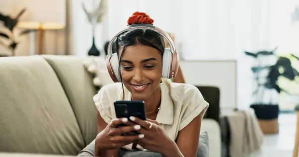 Smartphone, music headphones and happy woman on sofa in home, listening to audio or video app to relax. Phone, sound and Indian person on radio or typing on social media in living room on mobile tech.