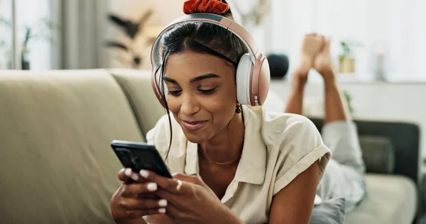 Phone, music headphones and happy woman on sofa in home, listen to audio and video app to relax. Smartphone, sound and Indian person on radio or typing on social media in living room on mobile tech.