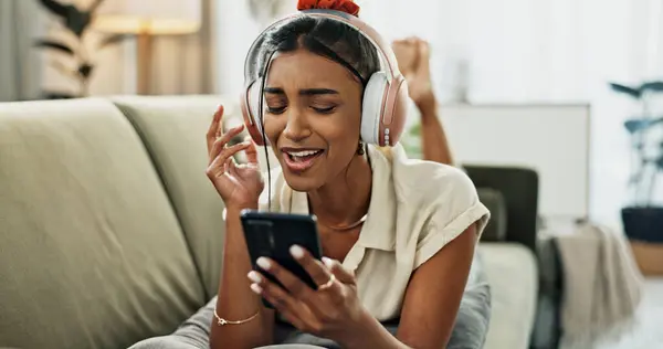 Phone, music and happy woman singing on sofa in home, dance and listening to audio. Smartphone, excited Indian person on headphones for radio, sound or freedom in living room on mobile app technology.