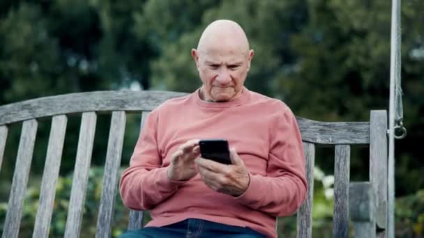 高齢者 ソーシャルメディア モバイルアプリやメッセージの読書のためのベンチで携帯電話と屋外 インターネットによるニュース 通知のためのシニア市民 スクロール — ストック動画