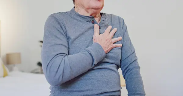 Hands, chest pain and closeup of elderly man in bedroom with injury, hurt or accident at nursing home. Sick, ill and zoom of senior male person in retirement with asthma or heart attack at house
