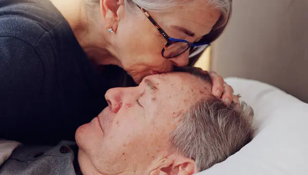 Senior woman kissing her husband on forehead with love, care and marriage in bed at home. Sick, recovery and elderly couple in retirement with illness in bedroom of hospice, nursing center or house