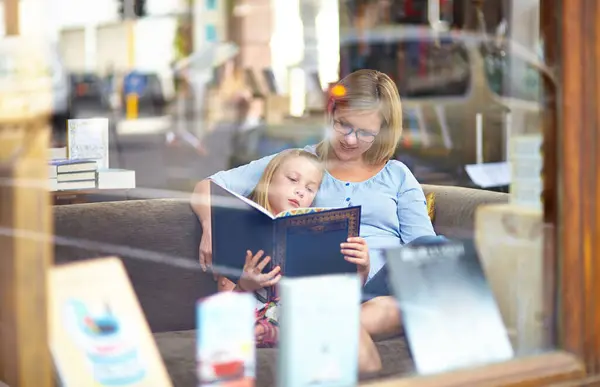 Reading, books and mother with child in bookshop window with smile, learning and relax with knowledge. Storytelling, happy mom and girl in library together with story, fantasy and education in window.
