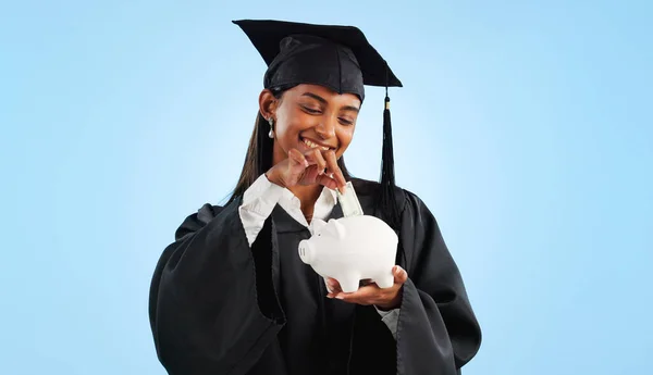 Woman, graduation and piggy bank in studio, saving and cash for investing, future or goals by blue background. Student girl, banking and money with thinking, decision and choice for financial plan.
