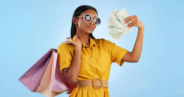 Woman, shopping bag and money fan, wealth and commerce with customer in sunglasses on blue background. Cash, financial freedom and retail, fashion and product choice, rich and shop discount in studio.