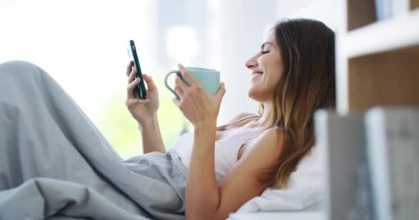 面白いソーシャルメディアの通知を読んだり チャットしたり 自宅でモバイルアプリをスクロールしたりするためにベッドで電話で笑う女性 スマートフォン ドリンク 笑顔の寝室でミーム 連絡先 ネットワークを検索する — ストック動画