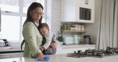 .Anne, bebek ve evde uyuyan bir çocuk ve ev işleri ile mutfak temizliği. Bir aile evinde büyümek ve gelişmek için küçük bir çocuğu tutarken tezgahı silmek için bez taşıyan bir kadın.