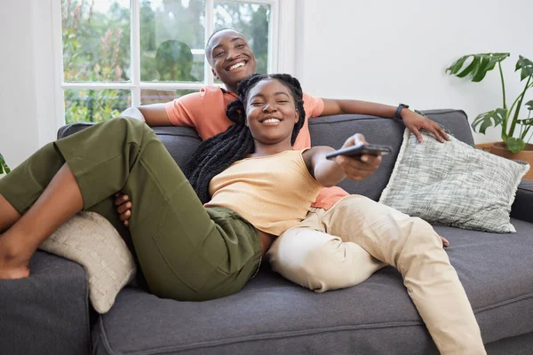 Young african american couple changing channels on remote and watching television together on sofa at home. Girlfriend relaxing on boyfriends lap while enjoying entertainment shows, series and movies.