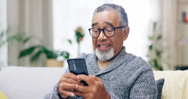 Senior man, happy and phone on couch for texting, reading or thinking for contact in home living room. Elderly person, smartphone and smile for notification, social media app and relax on lounge sofa.