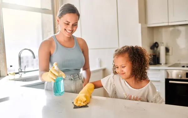 Little girl helping her mother with household chores at home. Happy mom and daughter wearing gloves while spraying and scrubbing the kitchen counter together. Kid learning to be responsible by doing .