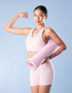 Mixed race fitness woman standing with her yoga mat and flexing her bicep in studio against a blue background. Beautiful young hispanic female athlete exercising or working out. Health and fitness.