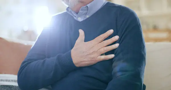 Hand, chest and heart attack with a senior man closeup on a sofa in the living room of his home during retirement. Healthcare, medical and cardiovascular disease with an elderly person in pain.