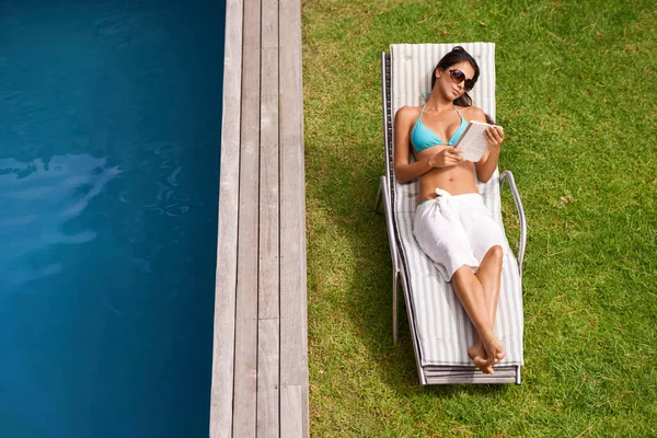 在吸收阳光的同时看书 一位年轻貌美的女子一边看书 一边在游泳池边休息 — 图库照片