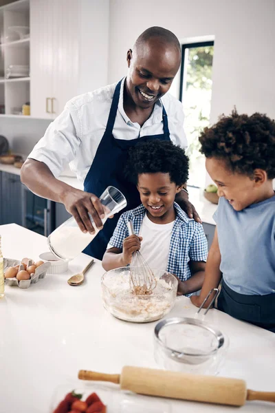 Adorable African American little boy with afro baking in the kitchen at home with his dad and brother. Cheerful Black man holding milk and mixing ingredients while bonding with his little boys.