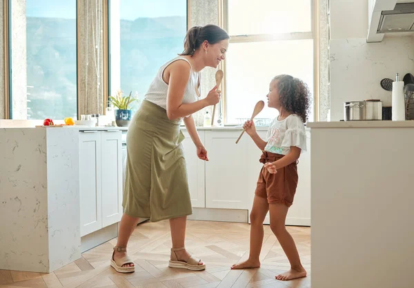 Mother and little daughter singing karaoke and dancing in the kitchen. Mixed race mom and child holding wooden spoons while having fun during a sing-off at home.