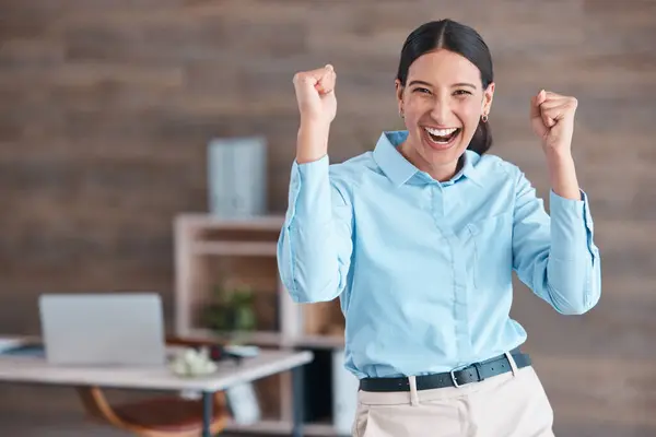 Young happy mixed race businesswoman standing and cheering with her fists alone in an office at work. One hispanic businesswoman celebrating success and victory at work.