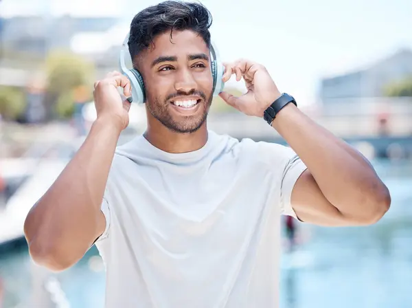 Happy young mixed race male athlete smiling while putting on his headphones before a run or jog and looking away outdoors. Fit sportsman listening to music while exercising.