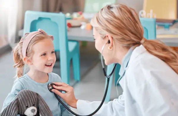 Young caucasian female doctor doing a checkup on a happy and cheerful little at a hospital. Carefree little girl smiling while holding a toy during a medical exam at daycare