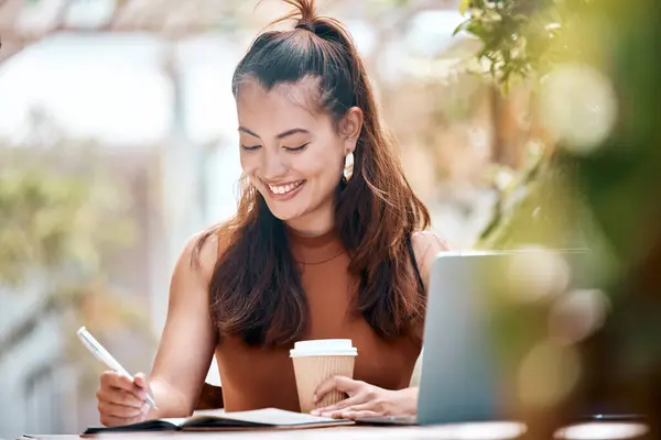 Young happy mixed race businesswoman writing in a notebook drinking coffee and working on a laptop sitting outside at a cafe. Hispanic female student smiling while studying at a restaurant.