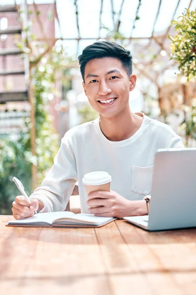 Young asian man or student writing notes and browsing on a laptop outside a cafe. One guy studying or working freelance at a restaurant. Doing research and planning online for blog, work or assignmen.