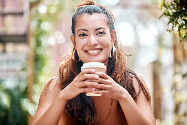 Happy young mixed race woman enjoying a cup of coffee on a break at a cafe in the city. One female only drinking a warm beverage in a disposable takeaway paper cup while relaxing outside a restaurant.