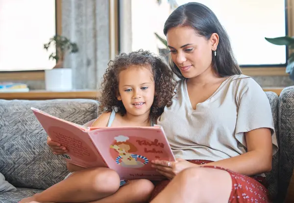 Loving hispanic mother and her little daughter sitting at home and reading a storybook together. Mother teaching little girl to read while sitting on the couch at home.