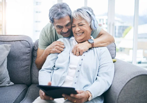 Senior, hug and couple with a tablet for a website, streaming music in home living room. Laughing, sofa and an elderly man and woman with a movie or funny video on technology or listening together.