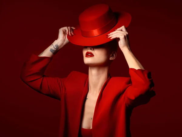 stock image Woman, red aesthetic and hat in studio with makeup art, thinking and mystery with couture, vision and background. Girl, model and high fashion with suit, mindset and facial paint for cosmetic beauty.