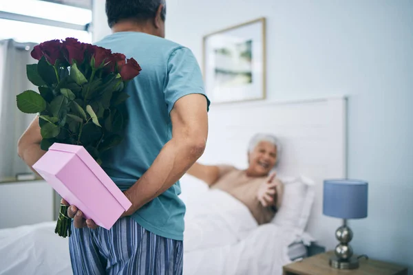 Gift, surprise and a senior couple on their anniversary in the bedroom of their house together for celebration in the morning. Flowers, birthday box and an old man giving his wife a present at home.