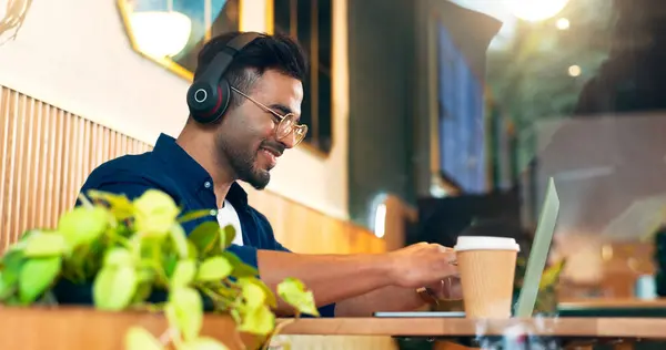 Happy man in cafe with headphones, typing on laptop and remote work, reading email and web in restaurant. Computer, freelancer smile and copywriter in coffee shop, listen to music and radio at table.