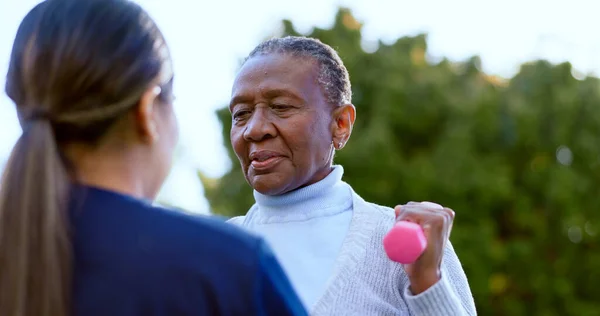 Dumbbell, fitness and a senior black woman with a nurse outdoor in a garden together for physiotherapy. Exercise, health or wellness with an elderly patient and medical person in the yard to workout.