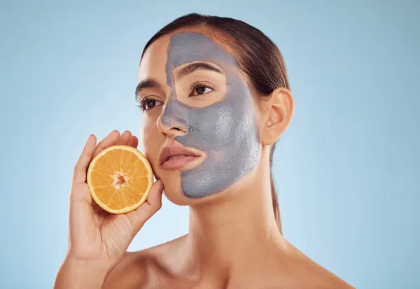 Woman, orange and face mask for skincare, vitamin C or natural nutrition against a blue studio background. Female person or model with healthy organic citrus fruit for beauty spa or facial treatment.