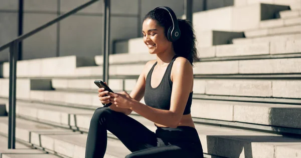 Runner girl, headphones and phone on stairs with music, smile and relax in city, workout and training. Woman, smartphone and sitting with audio streaming subscription with typing, social media or app.