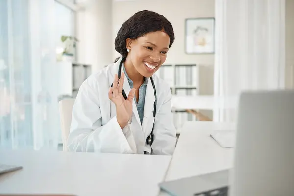 Video call, virtual medical meeting with a doctor waving, greeting and smiling with a laptop online working at a hospital. Professional healthcare worker doing telemedicine and giving advice.