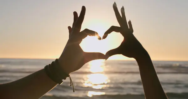 Love, hands and heart on beach with sunset, water background and horizon for vacation, travel or adventure. People, man and woman by ocean or sea with emoji for romance, care and relationship on trip.